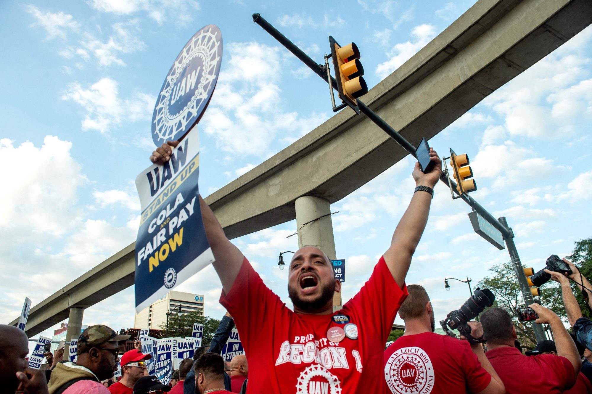 A UAW worker in a red shirt holds up a picket sign with both arms outstretched and a joyous expression on his face. Around him are other red-shirted UAW workers.